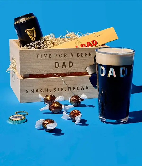 Fathers Day gift arrangement on a blue background featuring a wooden beer crate for dad, a pint of stout, chocolates, and a can of beer